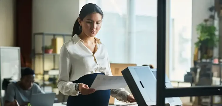 Businesswoman scanning documents at a copier in a busy office environment.